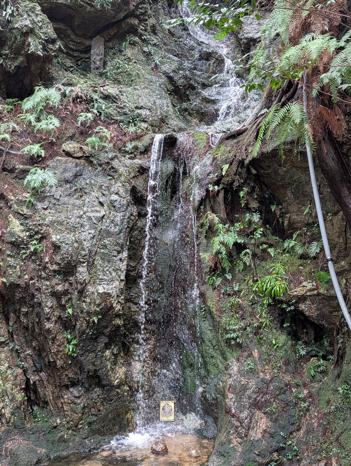 岩壁を細く落ちる滝。下の岩に小さな額